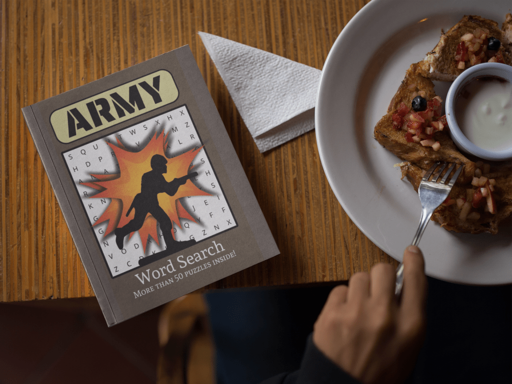 The Army Word Search Book is sitting on a table beside a person's hand that is holding a fork over a plate of food. This is breakfast with a relaxing word search book.