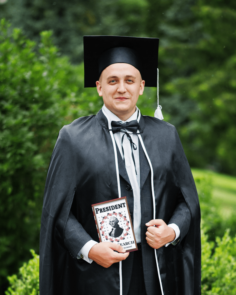 A graduate stands in a graduation gown with his hand holding a President Word Search Book. He looks proud.
