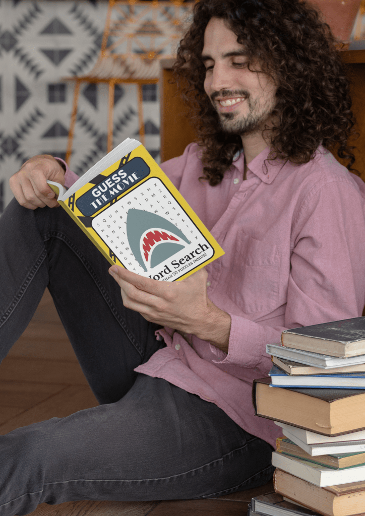 A happy man is sitting on the floor, leaning against a desk, with a stack of books beside him. He's reading a book called "Guess the Movie: Word Search Book" 