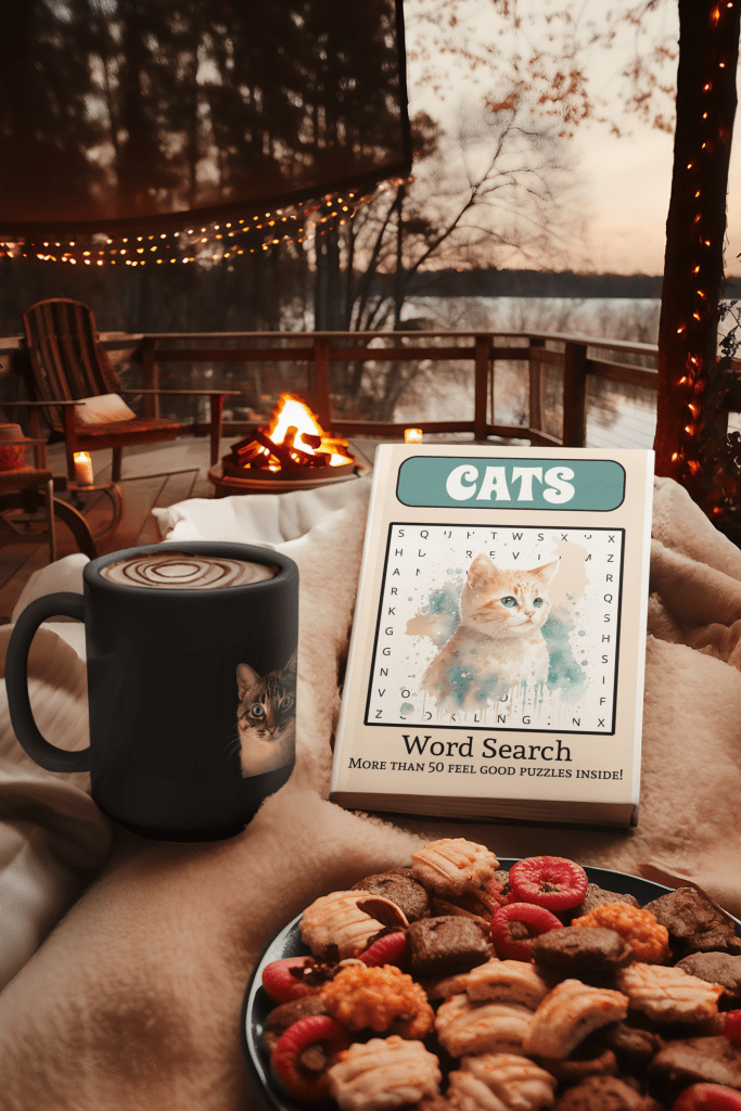 A cats word search book is on a table beside a hot mug of hot chocolate in front of a campfire.