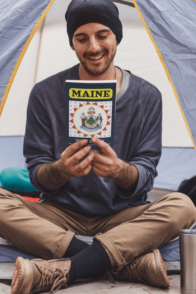 A young man sits cross legged in a tent, reading the back of the Maine Word Search Book and smiling.