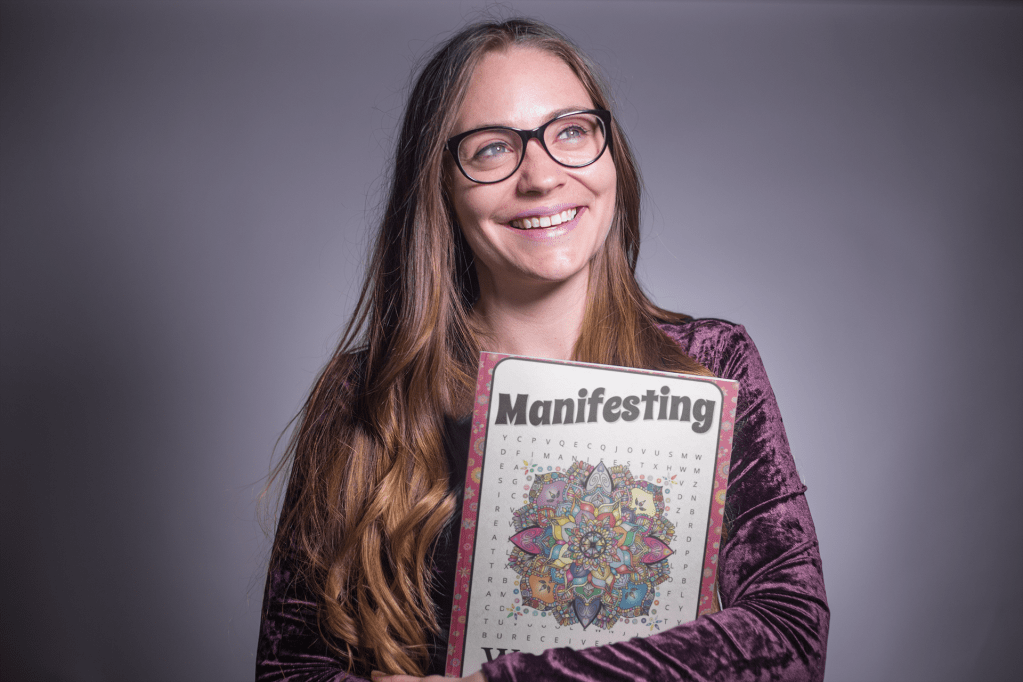 A young white woman with long brown hair and eye glasses is smiling while holding onto her Manifesting Word Search Book.