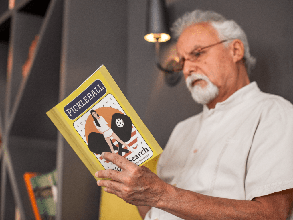 A senior man is reading a Pickleball Word Search Book. He's wearing glasses & appears to be concentrating.