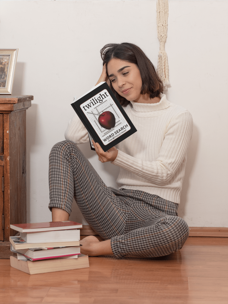 A young woman sits on the floor beside a stack of books. She's reading a Twilight Word Search Book with a small smile on her face.